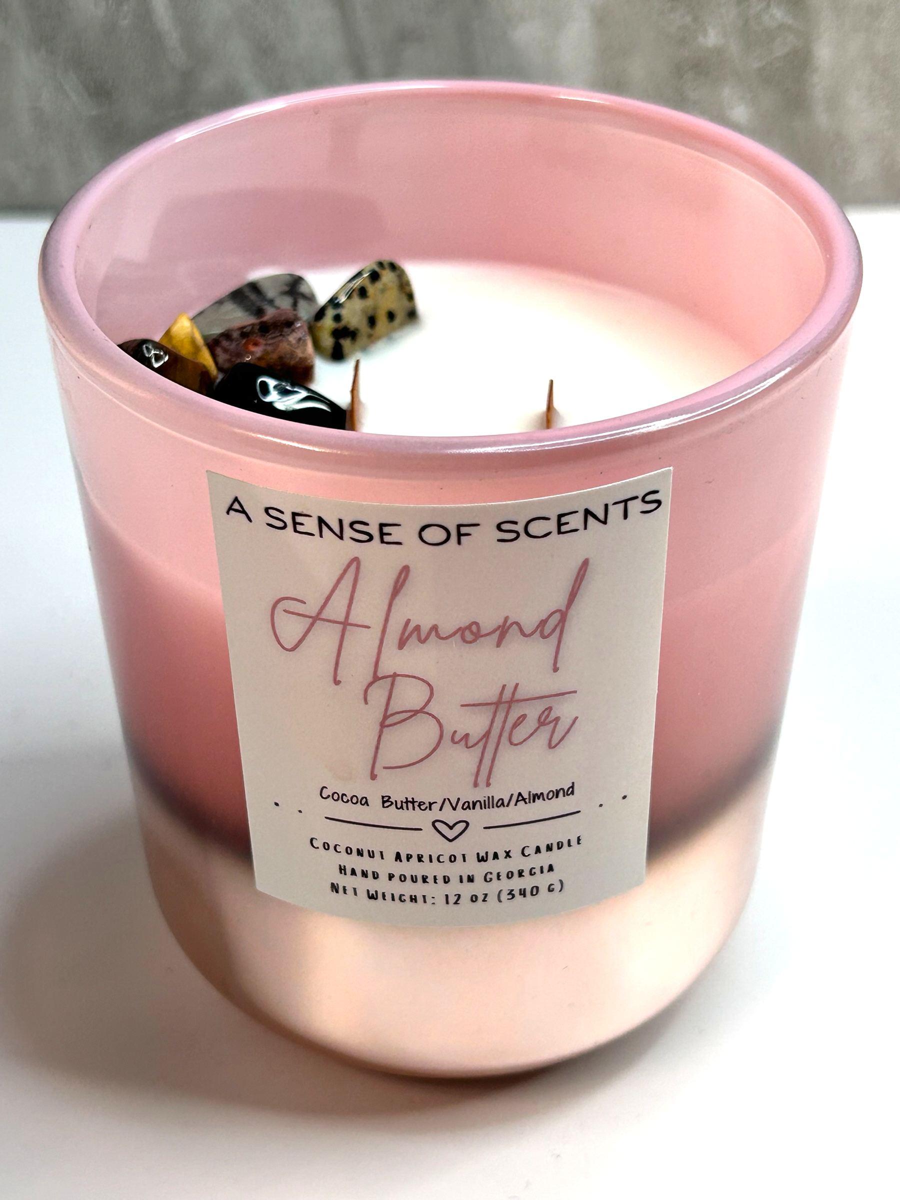 Pink candle labeled 'Almond Butter' from 'A Sense of Scents' on a white surface.