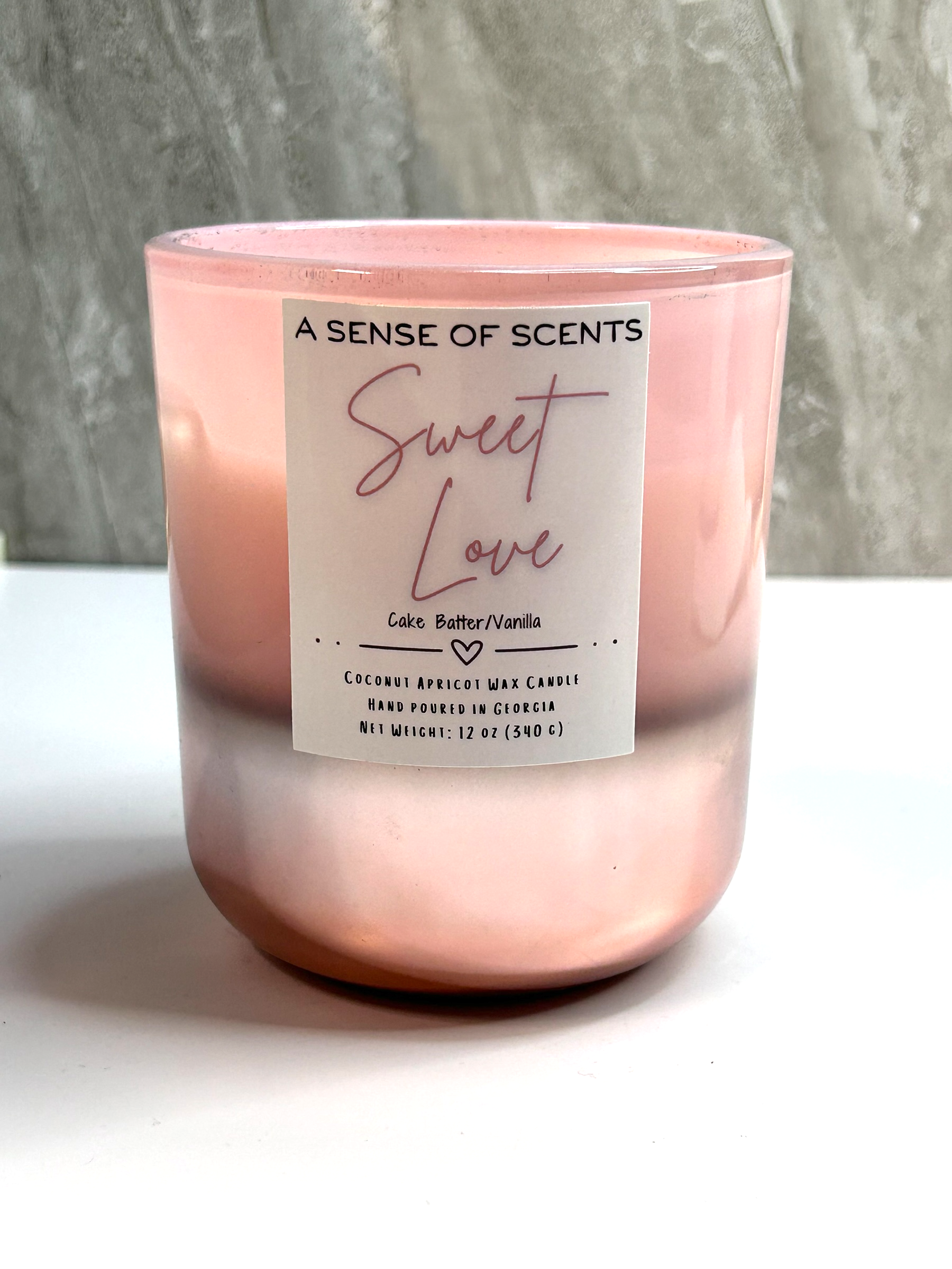 Pink candle labeled 'Sweet Love' by 'A Sense of Scents' on a white surface with a gray textured background.