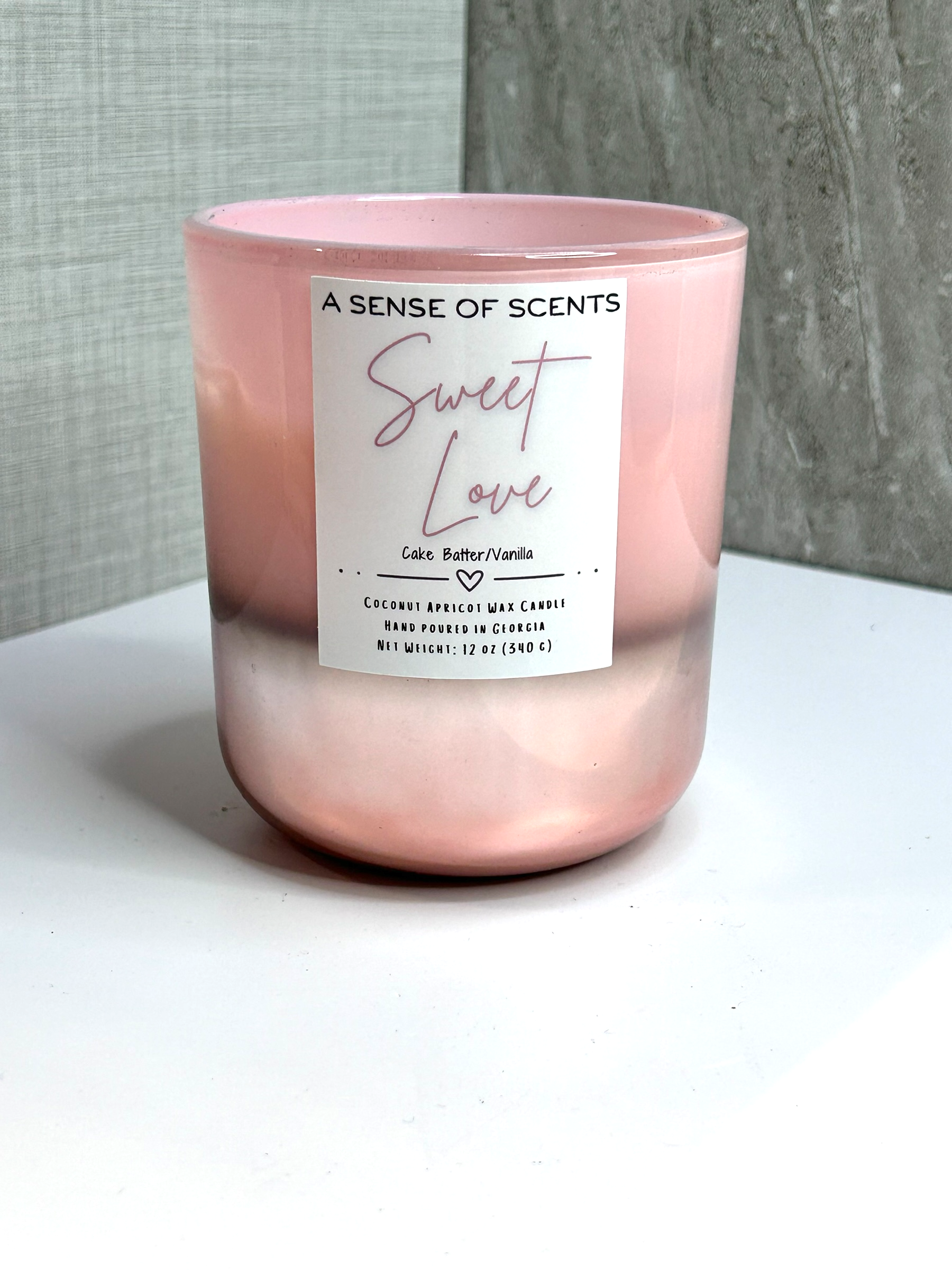 Pink candle labeled 'Sweet Love' by 'A Sense of Scents' on a white surface with a gray textured background.